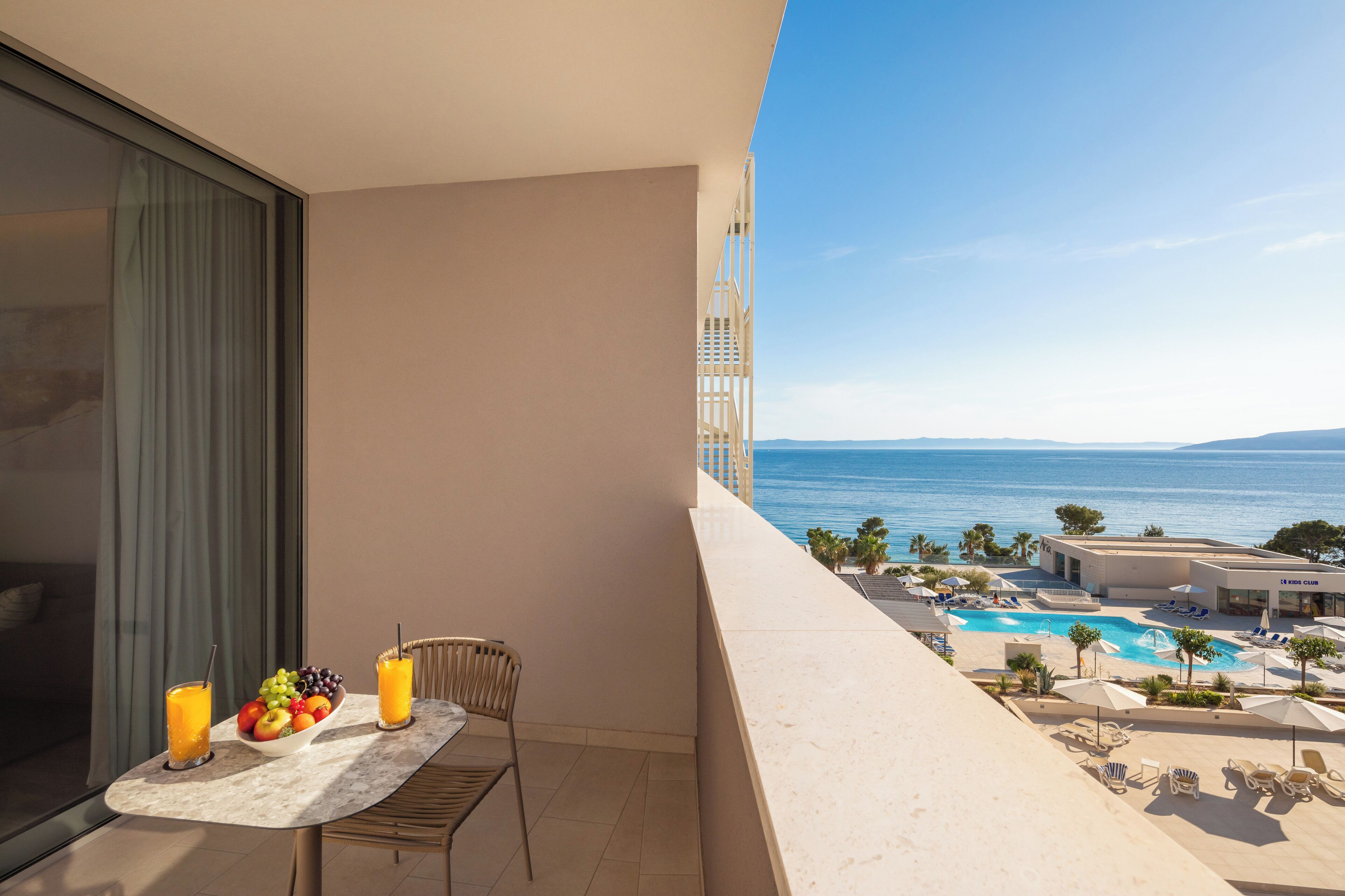 A balcony with a table set with fruit and drinks, overlooking a pool and ocean.