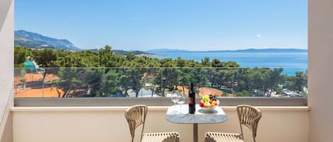 A balcony with two wicker chairs, a small round table, and a bottle of wine. The view includes a body of water and distant mountains.