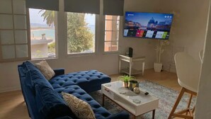 A living room with a blue sofa, a coffee table with decorative items, a television on the wall, and large windows offering a view of the outdoors. - Stylish Beachside. Parking.Views. FrontofBdg. (San Pedro)