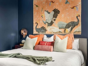 Une chambre à coucher avec un lit, des oreillers, une lampe fixée au mur et un tableau représentant des oiseaux.