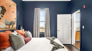 A bedroom with a large bed, a painting on the wall, a window with curtains, and a door leading to another room.