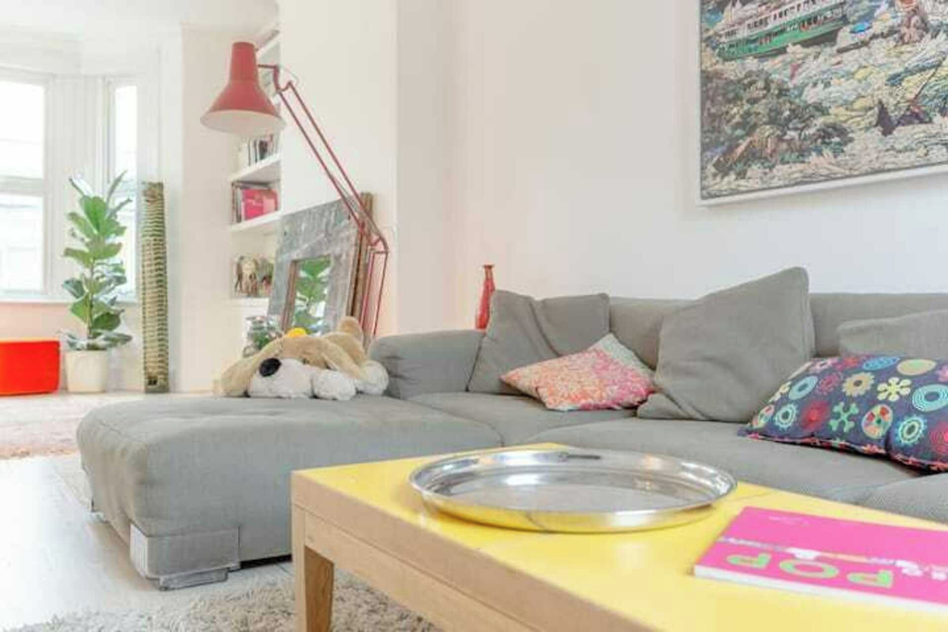 Een woonkamer met een grijze bank, een klein tafeltje met een metalen dienblad, een lamp en een kleurrijk kussen.