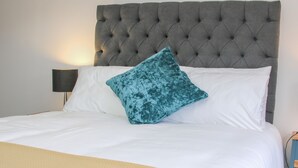 Una cama con cabecera capitonada, ropa de cama blanca y almohada color turquesa.