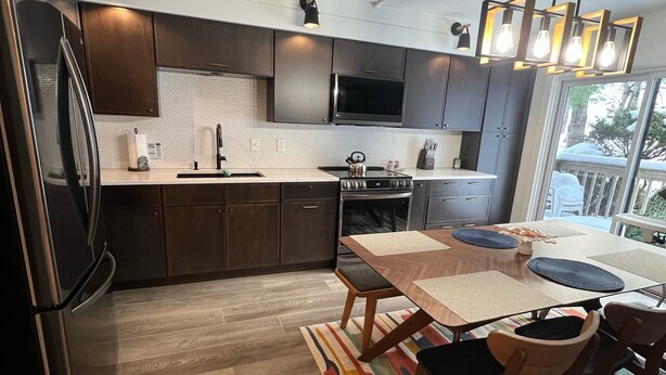 A modern kitchen with dark cabinets, a stainless steel refrigerator, a built-in oven, and a dining area with a wooden table and chairs. - Jay Peak's Premier Ski-In / Ski-Out Location-just steps from the trail (Jay)