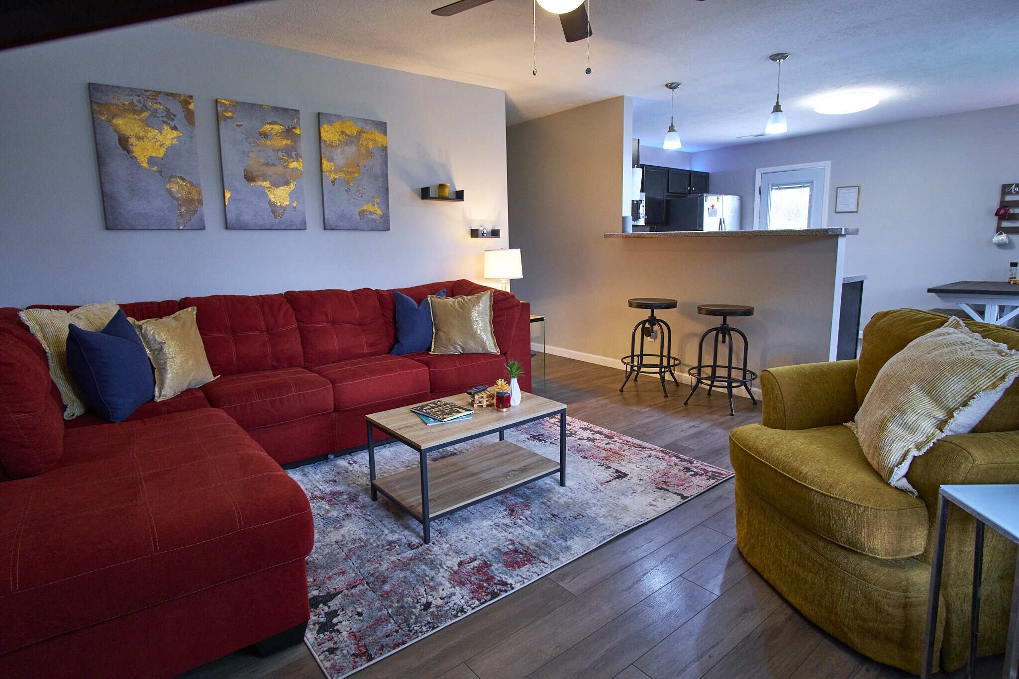 Una sala de estar con un sofá rojo, un sillón amarillo y una mesa de centro. Hay tres obras de arte colgadas en la pared que representan mapas.