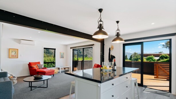 A modern kitchen with a large island, pendant lights, and a sliding glass door leading to a deck with a mountain view. - Views on Parry - Lake Hāwea Holiday Home (Lake Hawea)