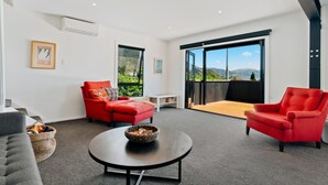 A modern living room with a red sofa, a grey couch, and a round coffee table. There is a large window with a view of mountains and a balcony with a wooden floor. - Views on Parry - Lake Hāwea Holiday Home (Lake Hawea)
