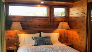 Un dormitorio de cabaña de madera con una cama grande, dos lámparas de noche y una ventana sobre la cama.