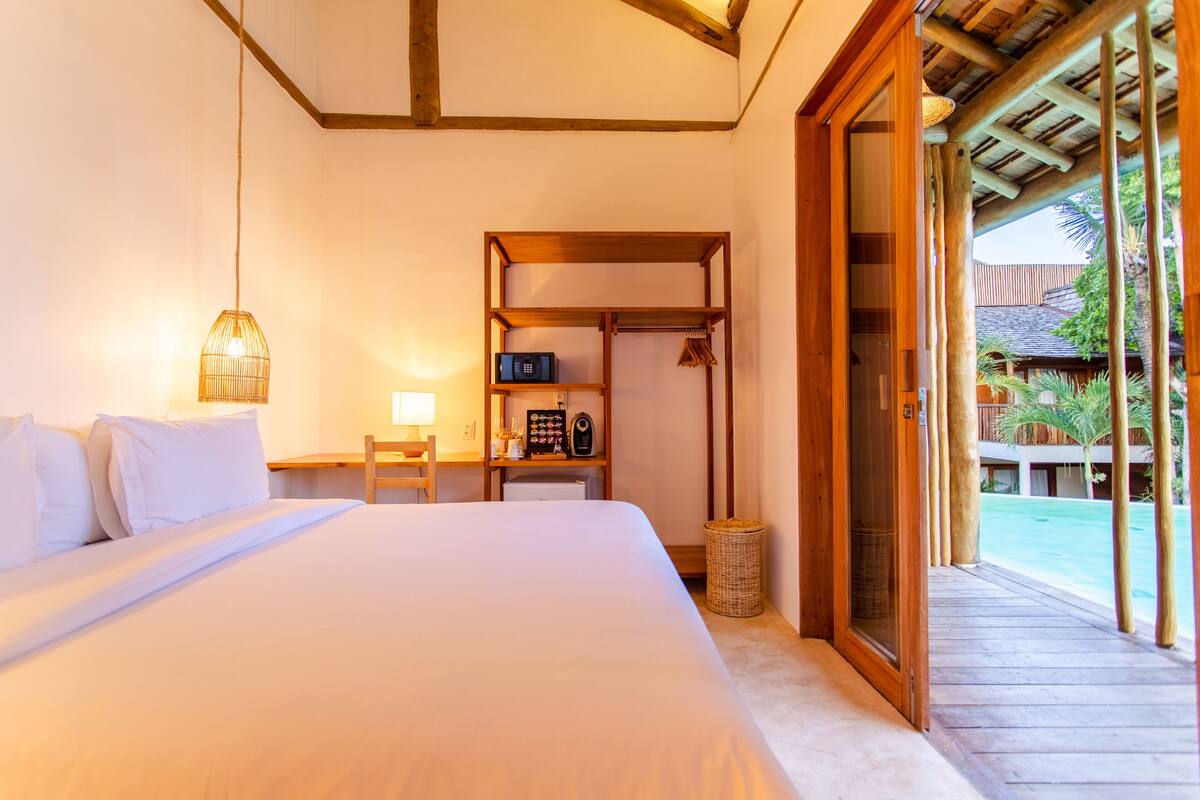 A bedroom with a large bed, a nightstand, a shelf with items, and a view of a pool and palm trees.