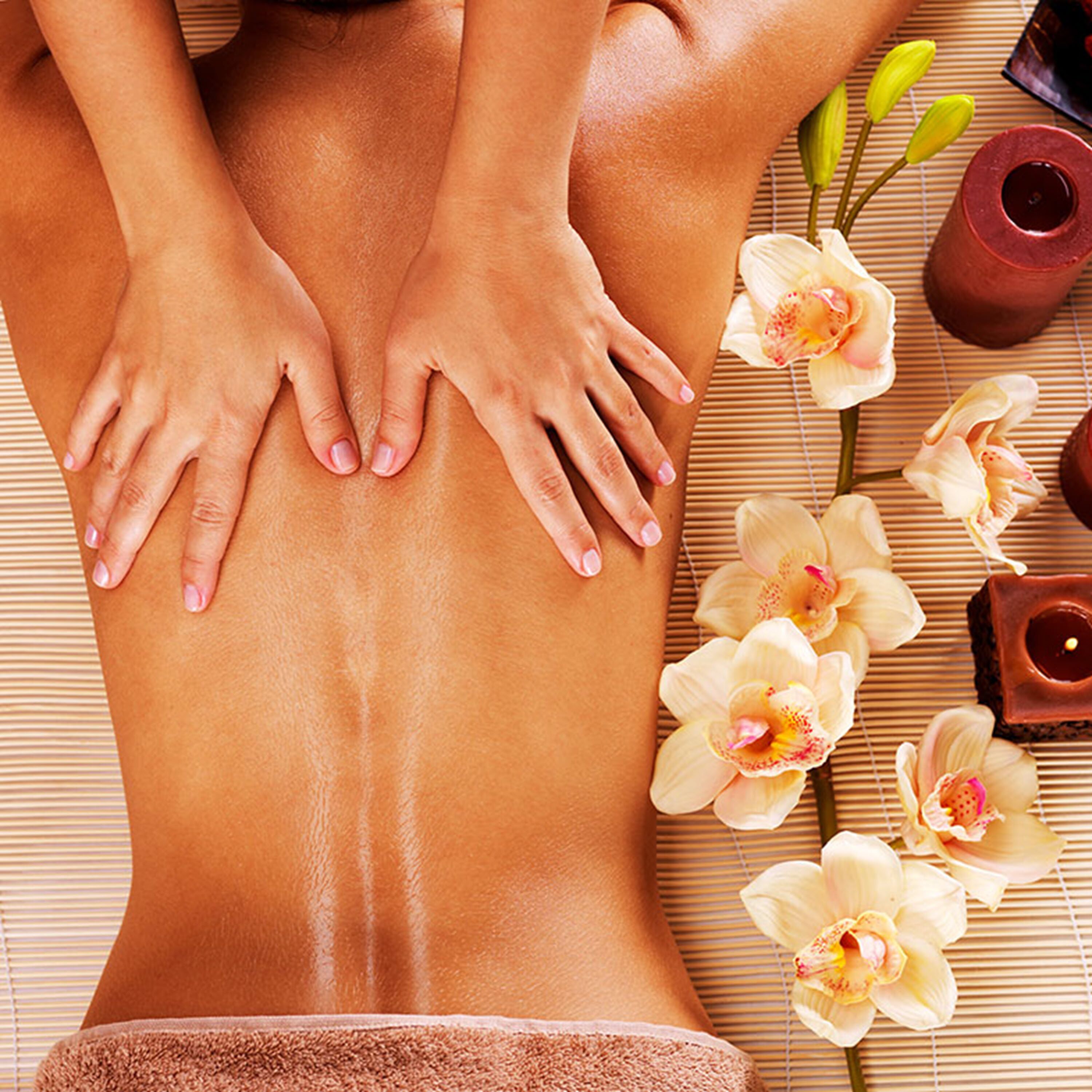 Une personne reçoit un massage, les mains de la masseuse étant posées sur son dos. Elle est entourée de fleurs et de bougies, le tout disposé sur un tapis en bambou.