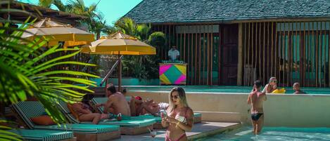 A pool area with lounge chairs and umbrellas, a wooden building with a tiled roof, and palm trees.