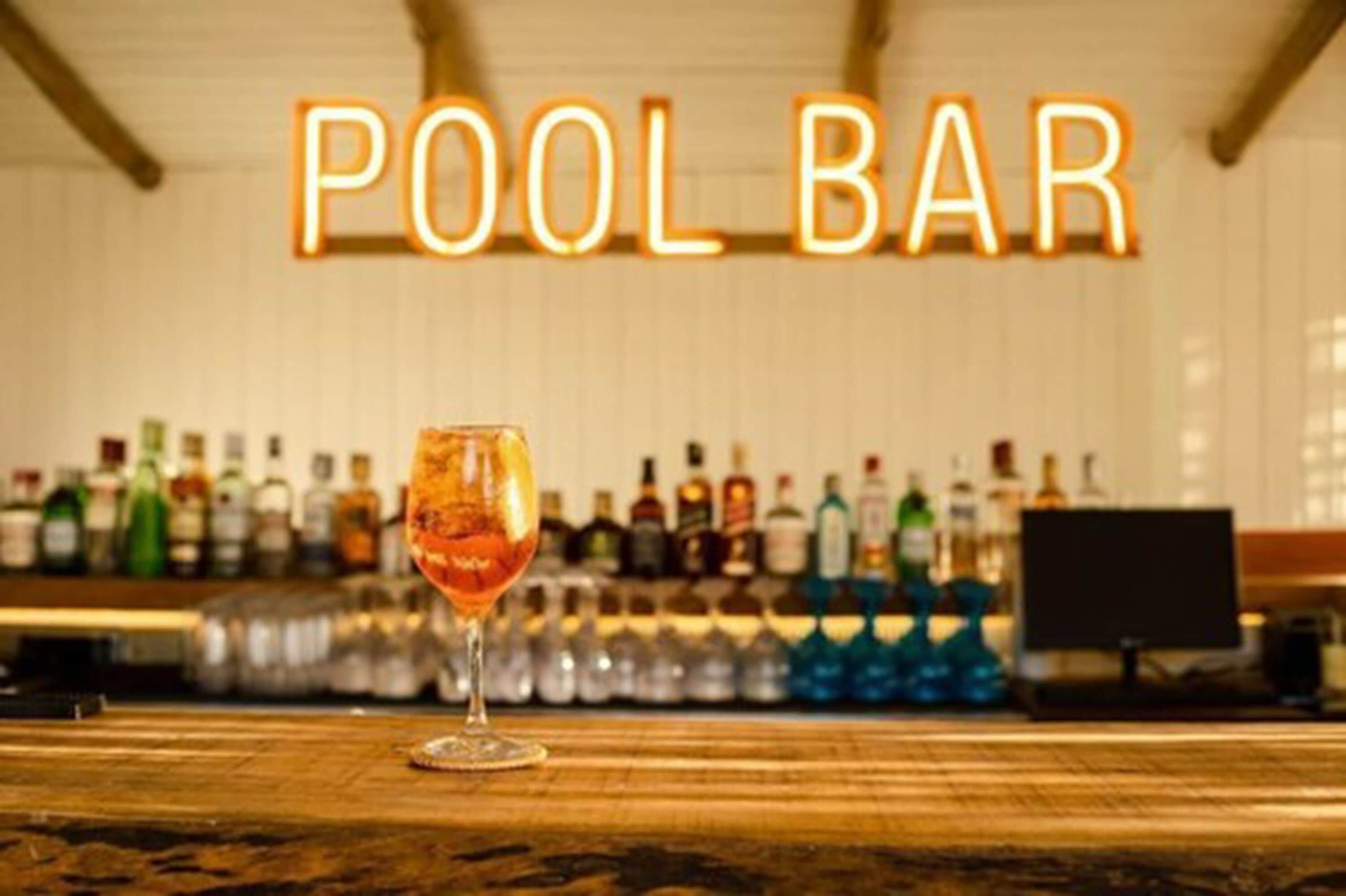 A bar with a neon sign reading "POOL BAR" and a glass of amber-colored drink on the counter.