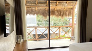 Een hotelkamer met een bed, een houten dressoir, een spiegel en uitzicht op een balkon met rieten dak.