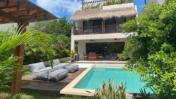 Een huis met twee verdiepingen, een zwembad, terras en tropische planten.