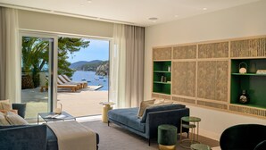 Un salon moderne doté d’une grande porte-fenêtre coulissante donnant sur un balcon avec vue sur la mer et les bateaux. On y trouve deux canapés, une table basse et une étagère verte adossée au mur.
