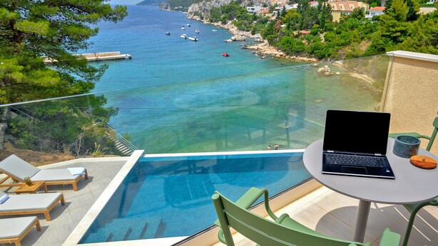 Un balcon avec un ordinateur portable, une petite table et une chaise, donnant sur une mer d’un bleu profond, parsemée de bateaux et bordée d’une côte rocheuse.