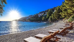 Une plage de galets avec des chaises longues en bois, offrant une vue dégagée sur la mer et sur une colline parsemée de bâtiments.