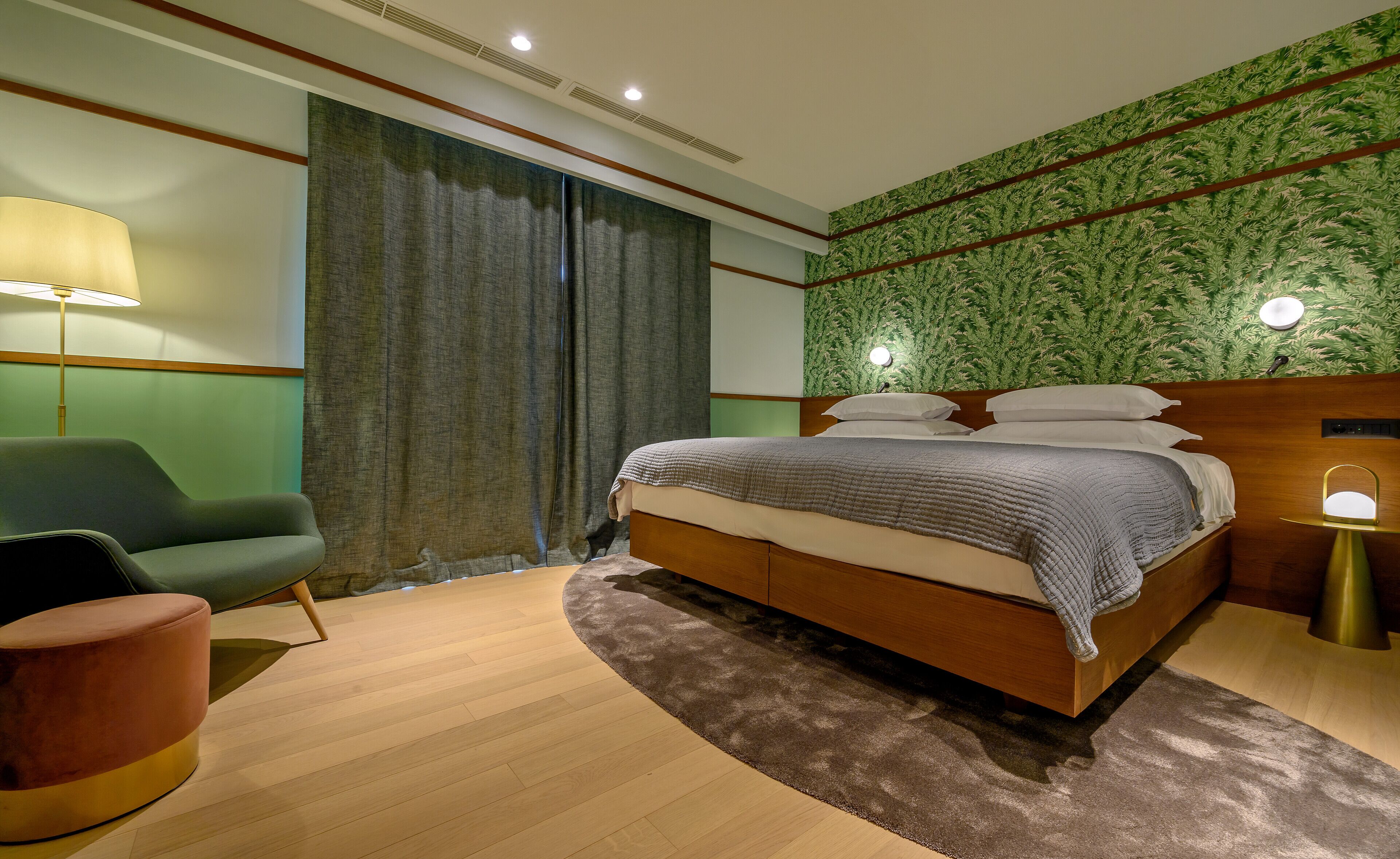 Un dormitorio moderno con una cama de madera, un sillón verde, un otoman redond y un ventanal con cortinas.