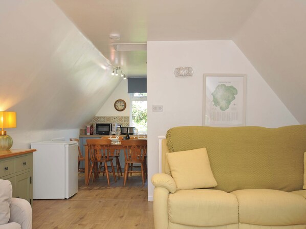A cozy living room with a yellow sofa, a dining area with wooden chairs, a kitchen with a microwave, and a clock on the wall. - 60 Valley Lodge (Gunnislake)