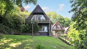 A two-story cabin with a deck, surrounded by lush greenery and a sloped lawn. - 60 Valley Lodge (Gunnislake)