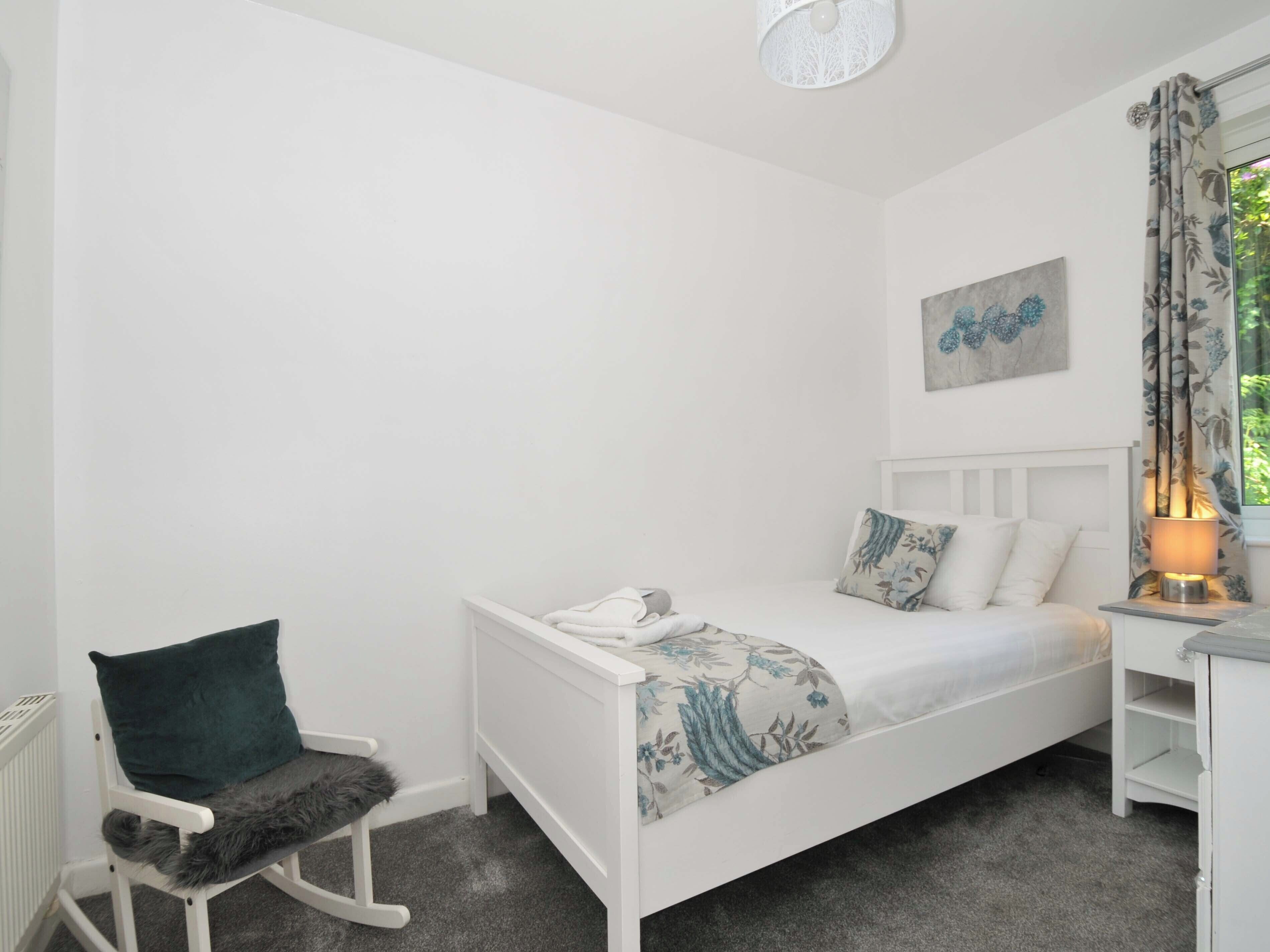 A bedroom with a white bed, a rocking chair, a bedside table with a lamp, and a window with curtains.