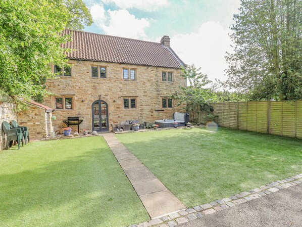A stone house with a tiled roof, a front door, and a small garden with a lawn and a paved path leading to the entrance.