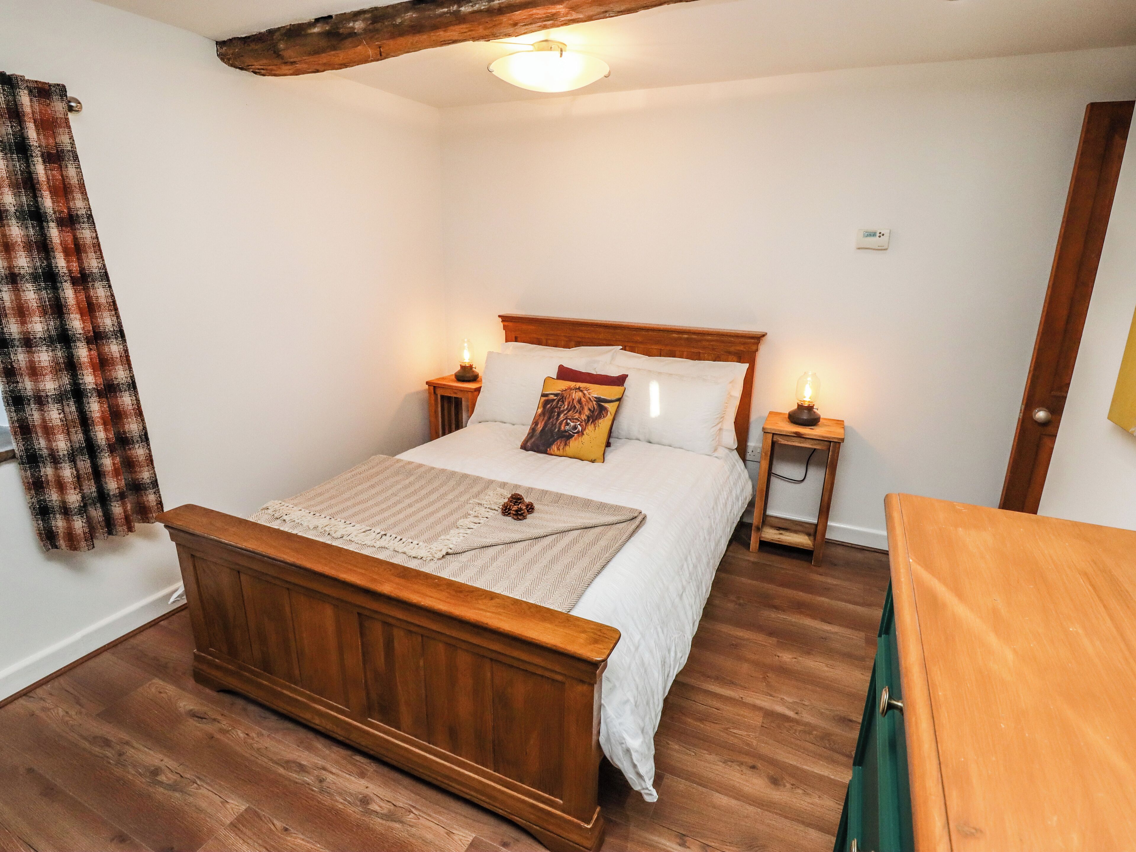 A bedroom with a wooden bed, white bedding, a wooden headboard, two bedside tables with lamps, a wooden dresser, and a plaid curtain.