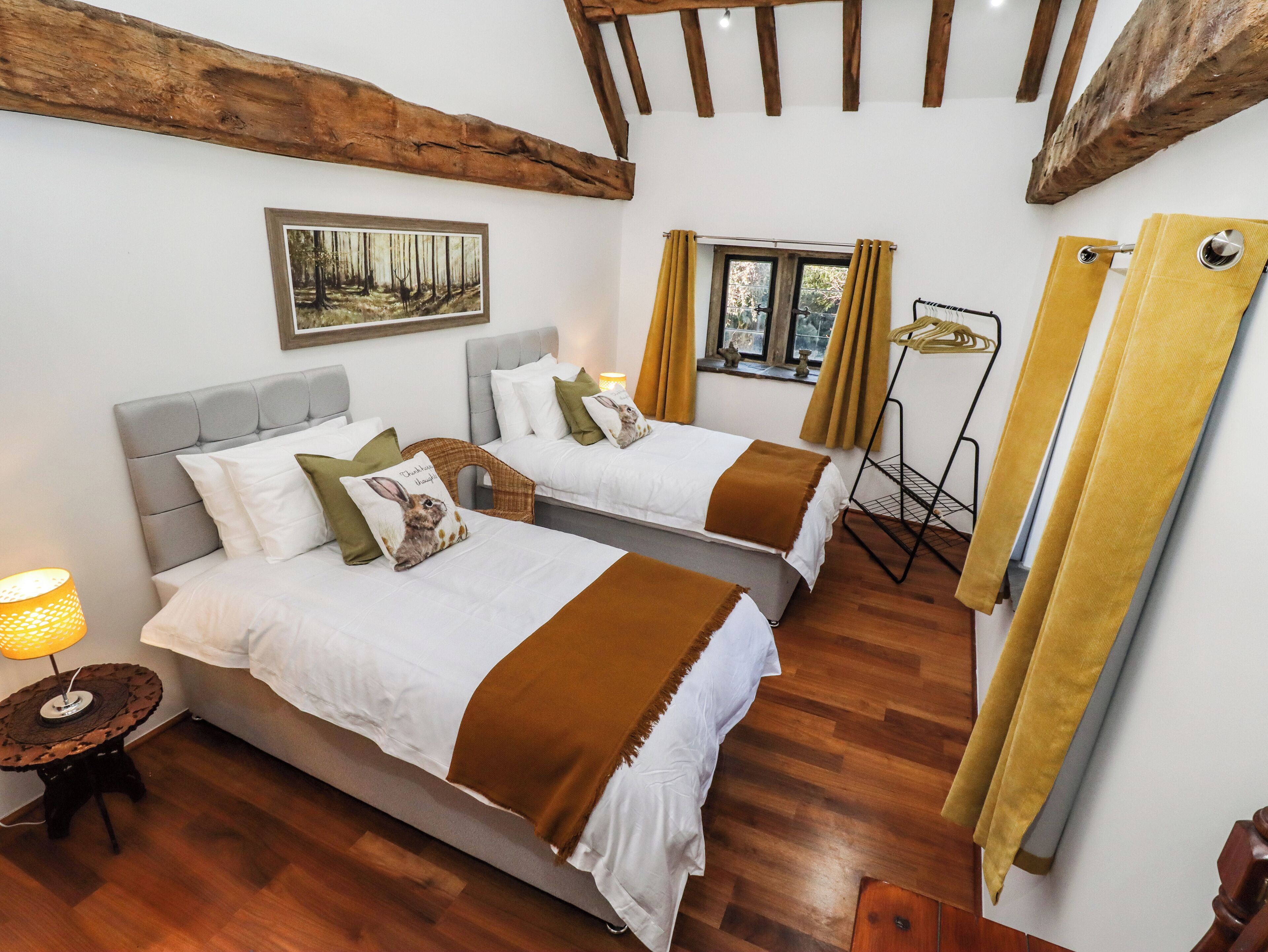 Ein Hotelzimmer mit zwei Betten, Holzboden, einem Fenster mit Vorhängen, einem Wandbild und einem kleinen Tisch mit Lampe.