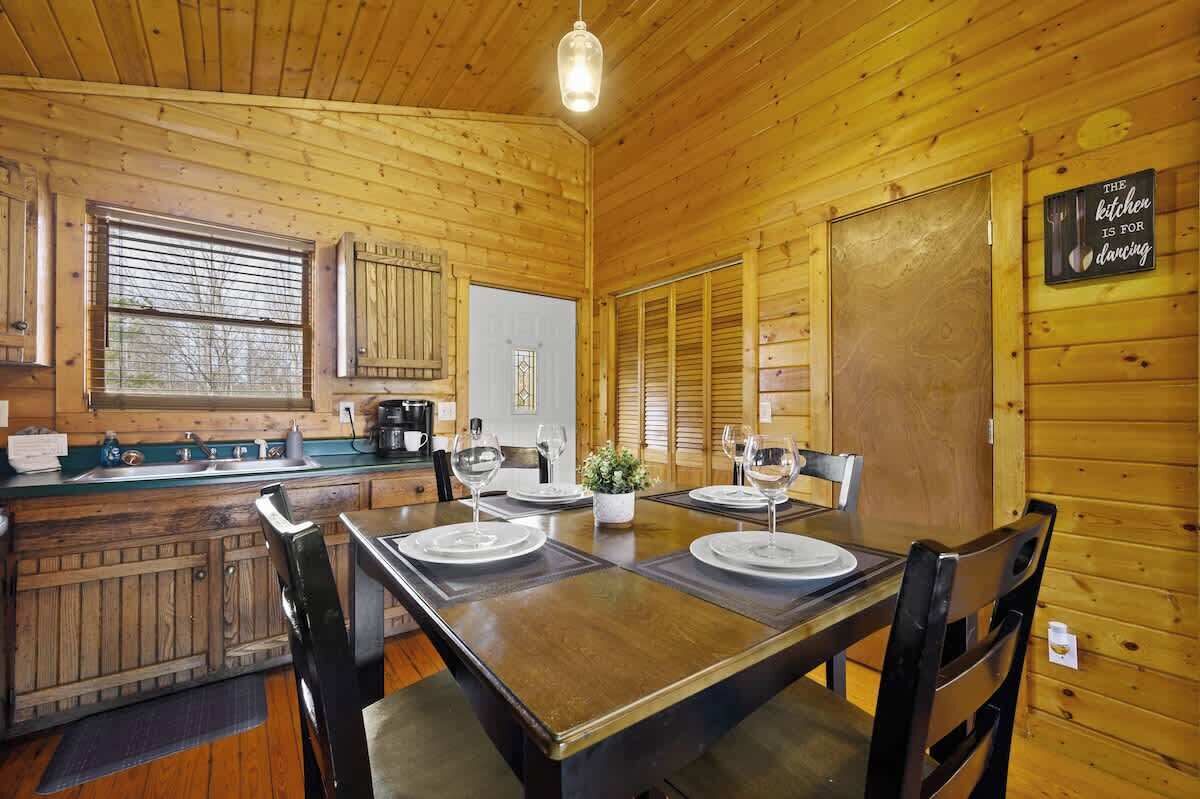 L’intérieur d’un chalet en bois comprend une salle à manger avec une table dressée pour quatre personnes, un coin cuisine et une pancarte accrochée au mur sur laquelle on peut lire : « La cuisine est faite pour danser. »