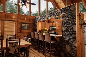 L'intérieur d'une cabane comprend une table à manger en bois, des chaises, un coin cuisine avec des murs en pierre et un ventilateur de plafond.