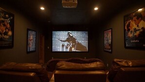 Una stanza buia con un grande schermo che mostra un film, poltrone in pelle e manifesti cinematografici alle pareti.