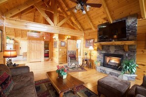 L’intérieur d’un chalet en bois, avec une cheminée en pierre, un téléviseur fixé au-dessus et un ventilateur de plafond.