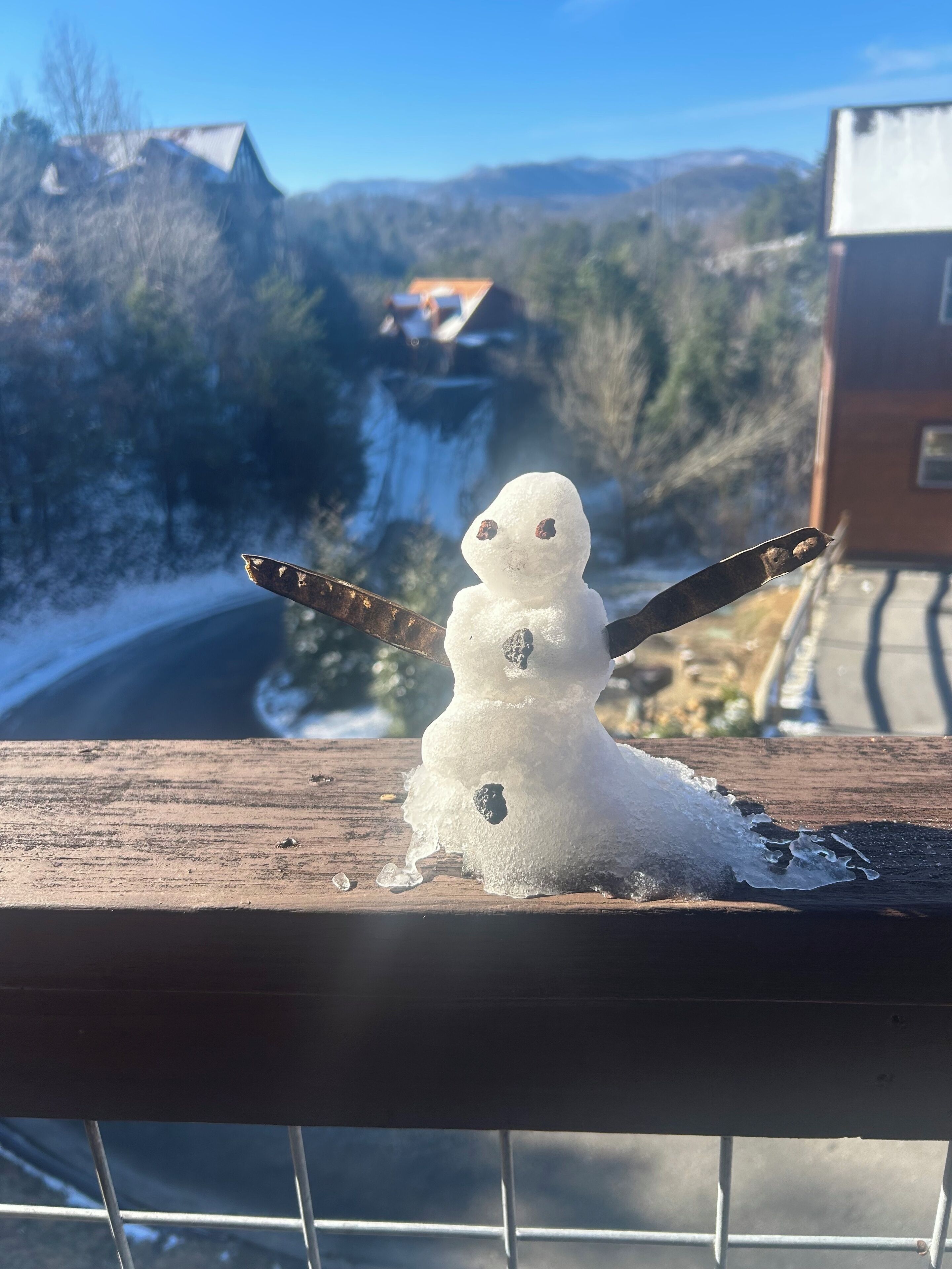 A snowman with a hat and a stick, placed on a wooden surface with a metal railing in the foreground.