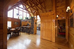 Une cabane en bois spacieuse comprenant un coin repas, une cuisine et un grand espace ouvert avec un plafond haut et des poutres apparentes.