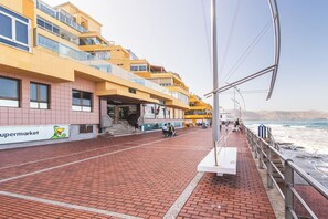 A coastal promenade with a modern building featuring a supermarket sign, a pedestrian walkway, and a view of the sea.