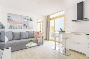 A modern living room with a grey sofa, a small round table with a plant, and a kitchen area with white cabinets and a black range hood.