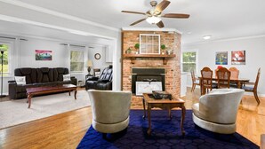 A living room with a fireplace, a ceiling fan, a sofa, and a dining area. - Modern Charm & Garden View sleeps 6 (Greensboro)