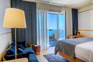A modern hotel room with a large bed, a sofa, a lamp, and a view of the sea through a sliding glass door.