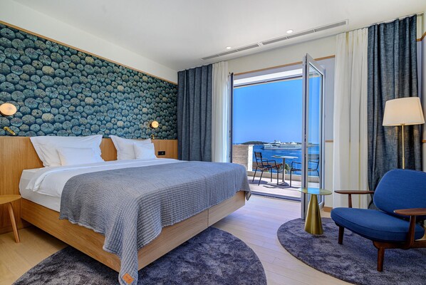 Une chambre d’hôtel moderne dotée d’un grand lit, d’un balcon avec vue sur la mer et d’un coin salon comprenant une chaise et une table.
