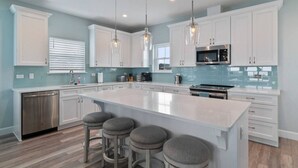 A modern kitchen with white cabinets, a large island with bar stools, stainless steel appliances, and pendant lighting. - Just Beachy: Stunning 4BR Oceanfront Home + Private Pool (St. Augustine)