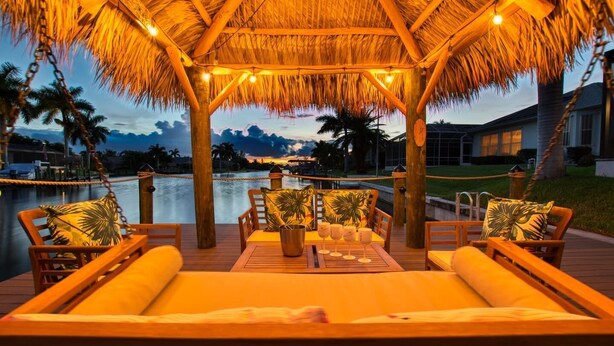 A thatched-roof outdoor seating area with wooden furniture and a view of a body of water and palm trees.