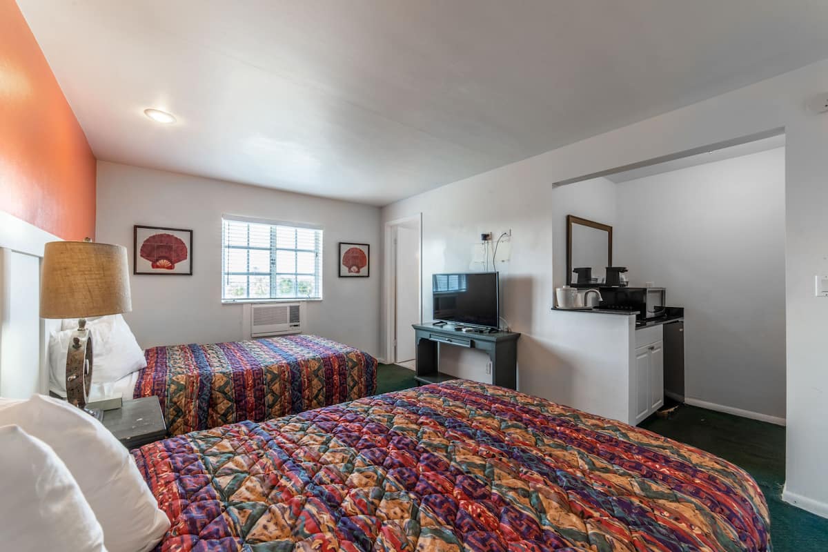 A hotel room with two beds, a desk with a TV, a kitchenette, and a window with blinds.