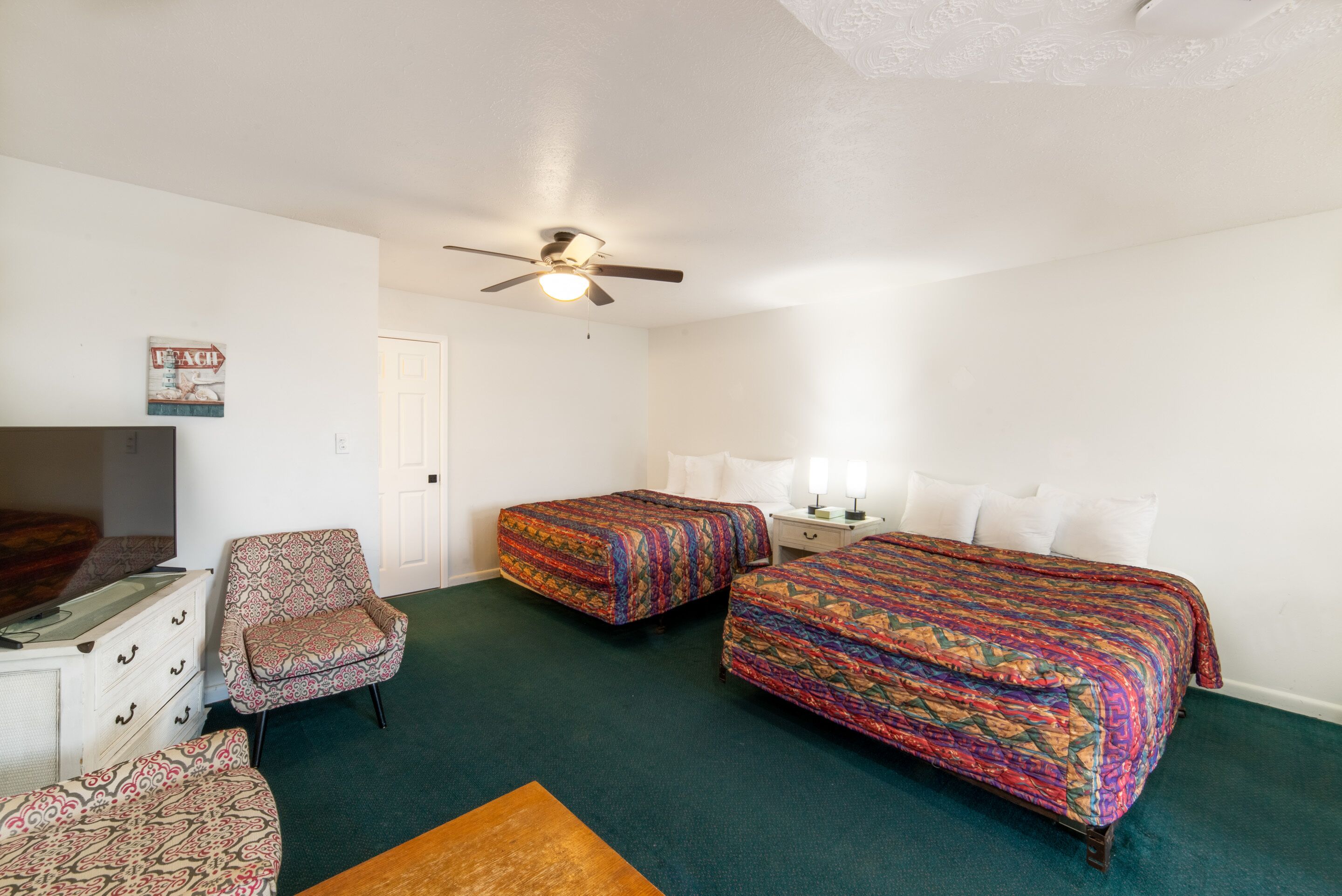 Une chambre d’hôtel avec deux lits, une chaise, une commode, un ventilateur de plafond et une porte.
