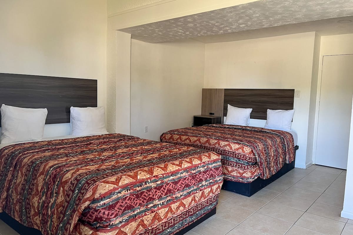A hotel room with two beds, each with a patterned bedspread and white pillows. The room has a simple, clean design with a tiled floor and a plain ceiling.