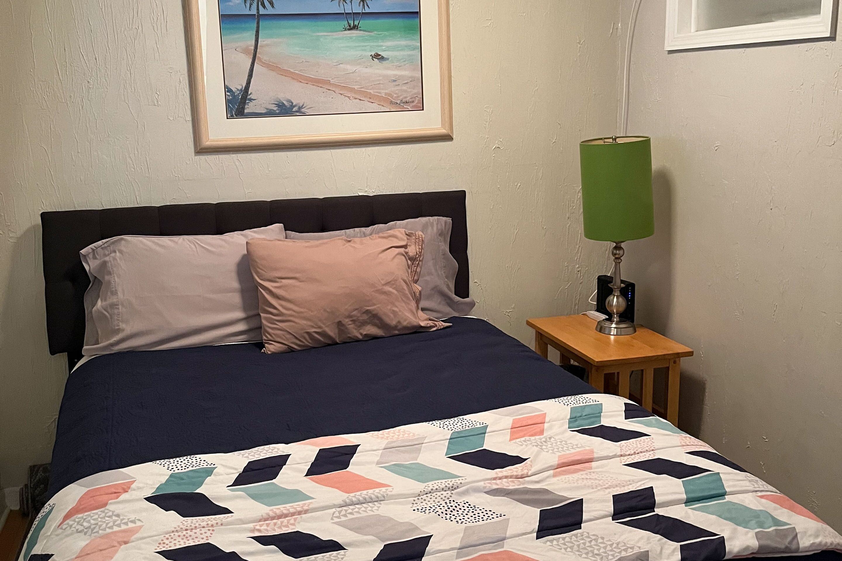 A bed with a dark blue bedspread and a multicolored quilt, a framed beach painting on the wall, and a green lamp on a wooden nightstand.