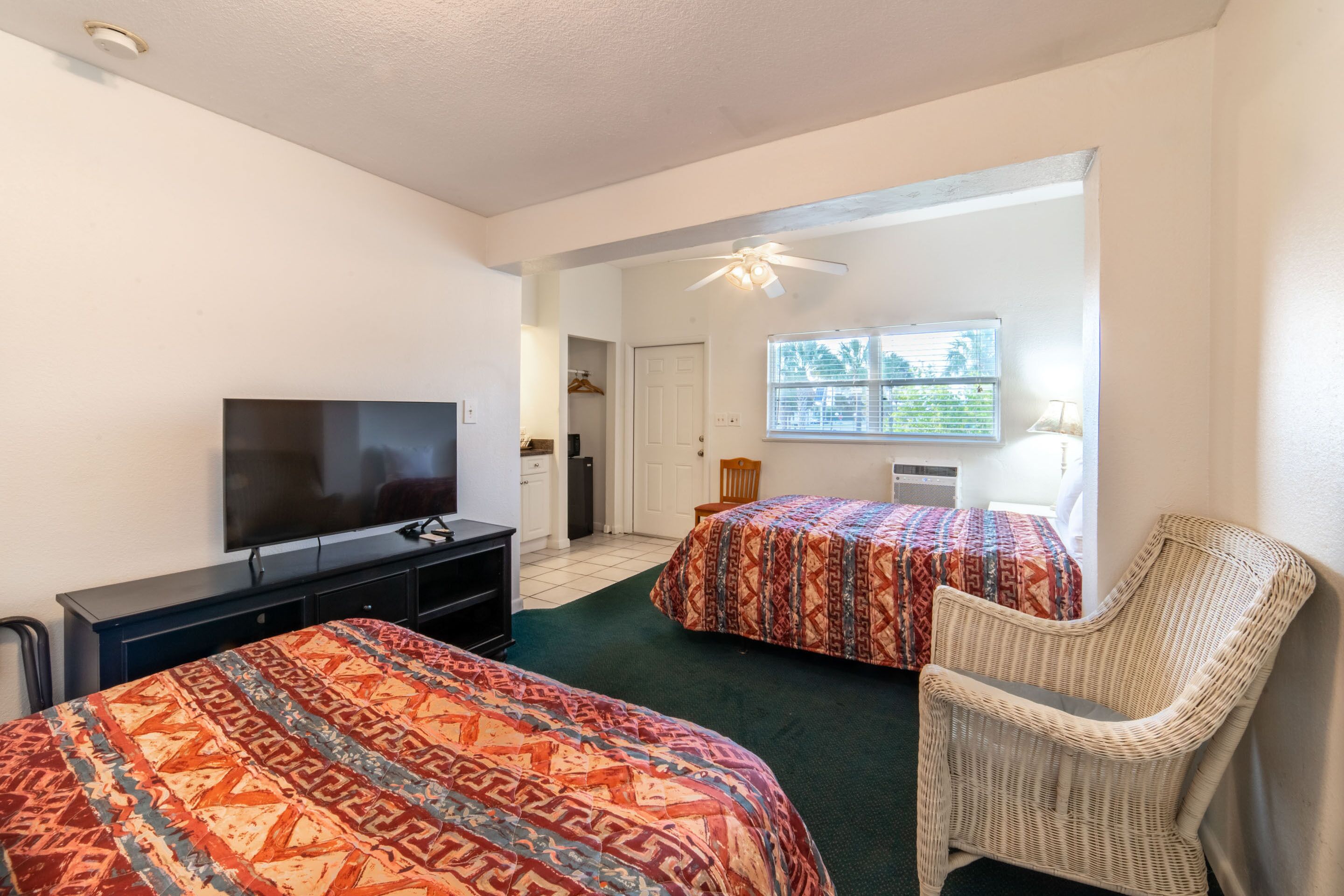 A hotel room with two beds, a television, a wicker chair, and a window with blinds.