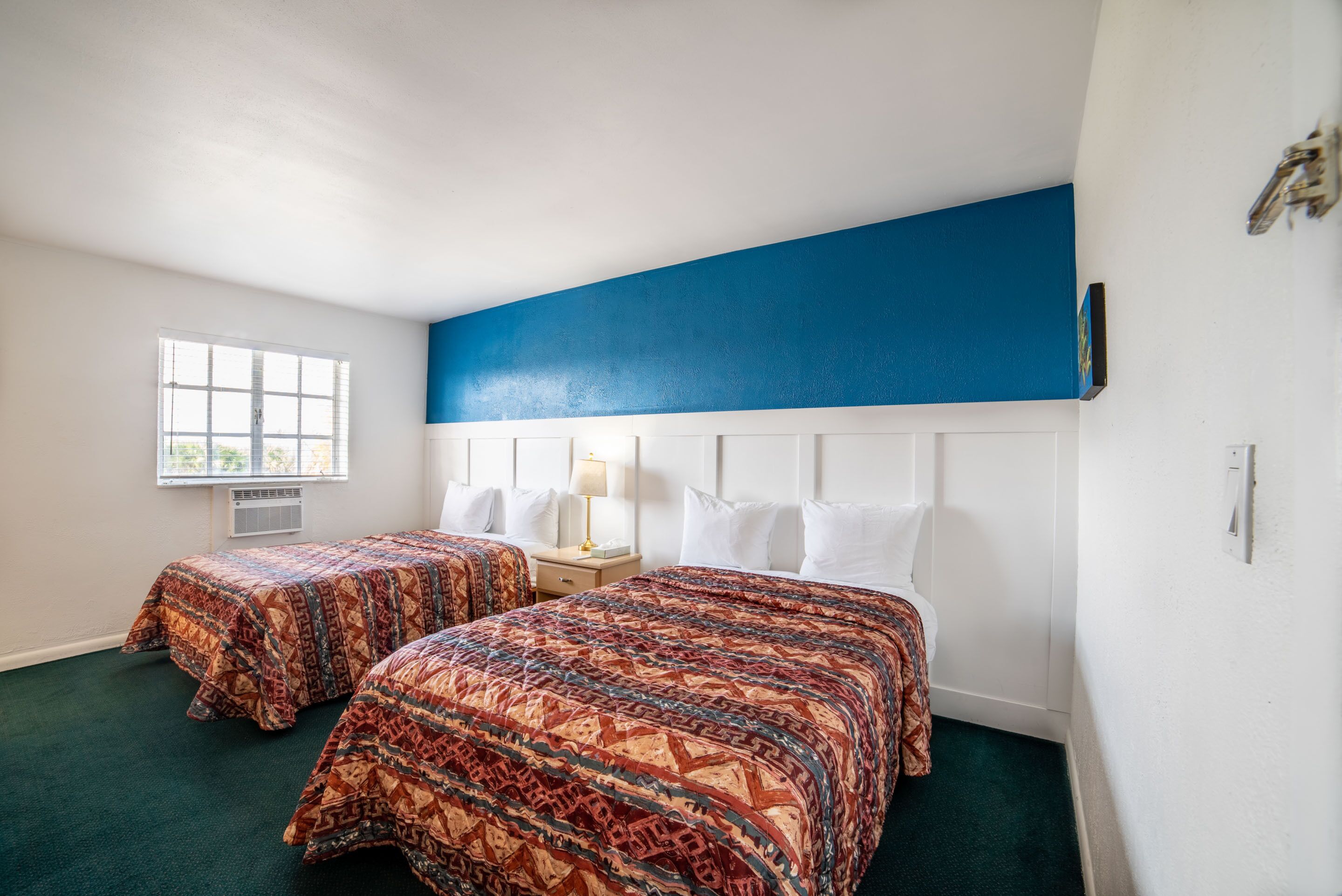 Una habitación de hotel con dos camas, una ventana, un cabecero blanco y una pared de acento azul.