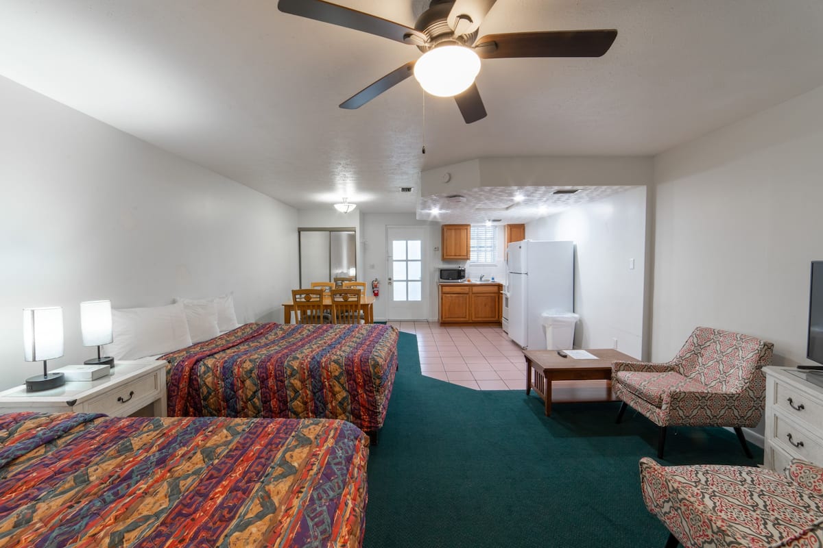 A hotel room with two beds, a desk, a chair, a TV, and a kitchenette in the background.
