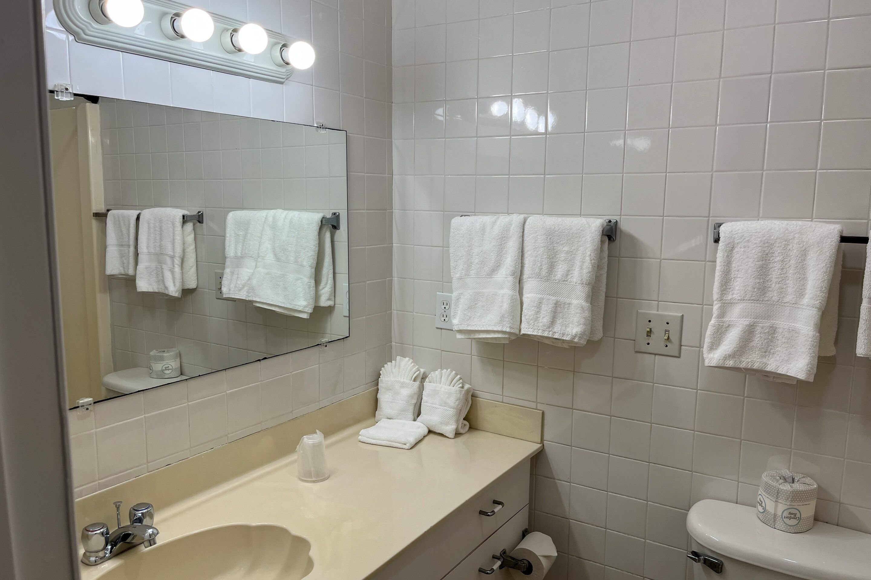 A bathroom with white tiles, a sink with a mirror above it, and multiple towels hanging on hooks.