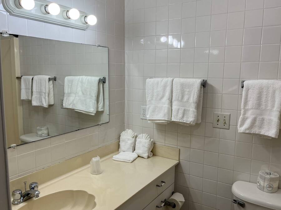 A bathroom with white tiles, a sink with a mirror above it, and multiple towels hanging on hooks.